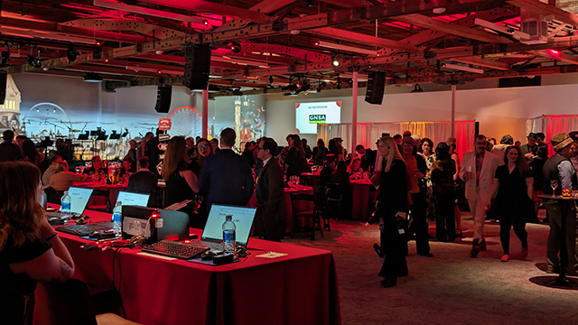 A person staffs a check-in table at a nonprofit gala. On the table are three open laptop computers. Guest mingle throughout the event space.