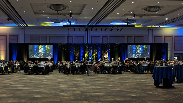A nonprofit gala in a large ballroom features to large screens on either side of the stage. Blue and gold lighting illuminate the black drape behind the stage and screens.