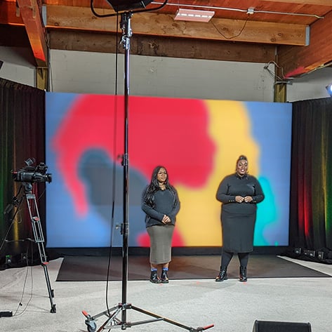 Two virtual event hosts stand in a production studio in front a video wall displaying a colorful, abstract image.