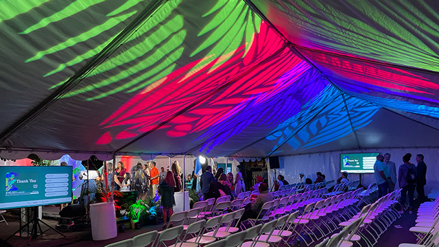Dynamic lighting adds drama to the inside top of a large event tent. White folding chairs are in rows facing the stage where people are gathered.