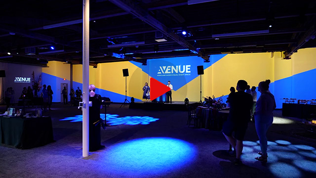 At AVENUE, the 85-foot projection wall  displays a blue and yellow graphic with the AVENUE logo. Two presenters are on stage and spotlights highlight different areas of the event venue.
