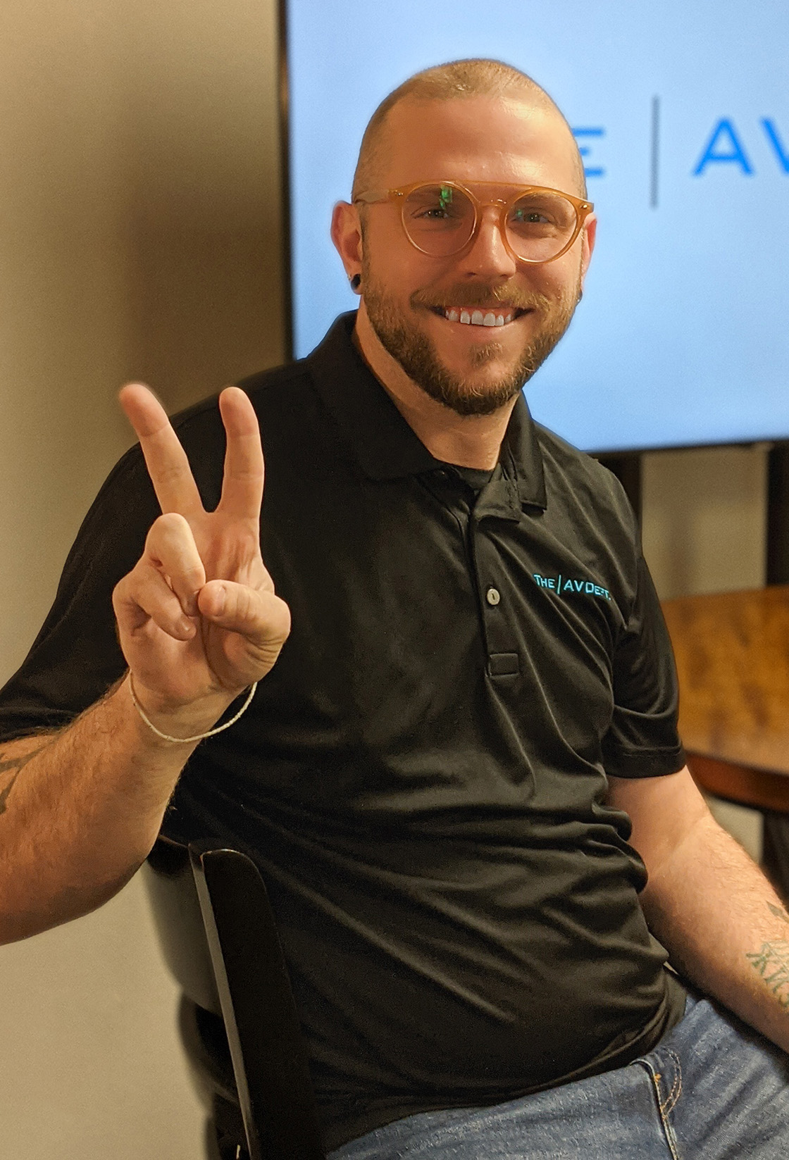 Devin Eaton, The AV Department Technical Events Planner hold up two fingers in a peace sign. He is sitting at a table in front of a large monitor that displays The AV Department logo
