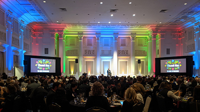 A rainbow of uplights in red, green, and blue light the perimeter of the Governor Ballroom at Sentinel Hotel. Two large screens on either side of the stage display a slide thanking sponsors of Virginia Garcia Memorial Foundation's ¡Prospera! 2023.