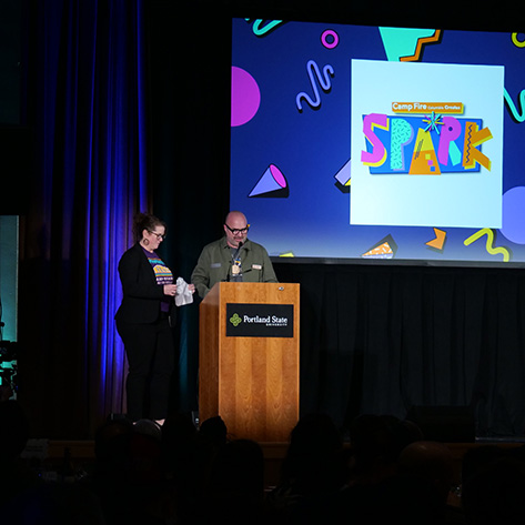 Two speakers are on stage between a podium and a large screen. On the screen is the logo of the hybrid event.