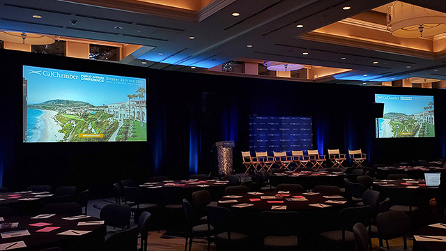A hotel ballroom is set for a conference with two large screens on either side of the stage. On the stage are 7 chairs for a panel discussion.