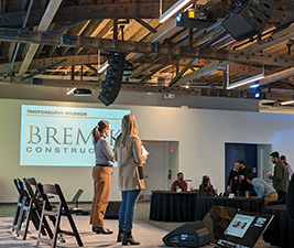 Two presenters speak to the audience from the stage. In front of the stage is a confidence monitor. Above the stage is a line array speaker sound system.