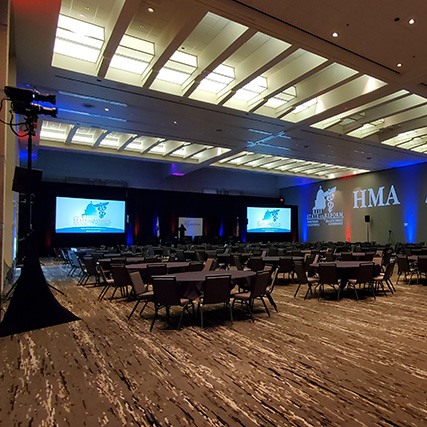A ballroom in the Sacramento Convention Center is set for the 2023 Northern Califronia State of Reform Health Policy conference. There are lighting trees, two screens, red and blut uplights and a stage in front of round tables with chairs
