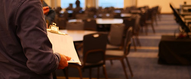 A techinical director holding a clipboard talks with a client in an event space. There are many tables and chairs in the background.