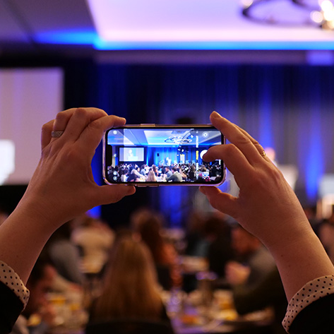 A conference attendee raises a cell phone high to take a picture of the keynote speaker