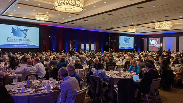 Hundreds of people sit at round tables in a hotel ballroom during a luncheon keynote address during a conference. Around the room are red and blue uplights. There are two large screens on either side of the stage.