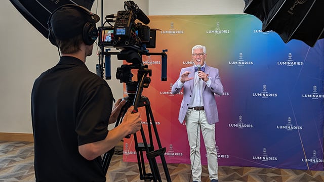 A videographer conducts an on-camera interview during an event. The guest is holding a microphone, looking toward a camera and stands in front of a step and repeat backdrop