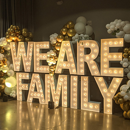Illuminated letters spelling We Are Family are surrounded by gold and white balloons