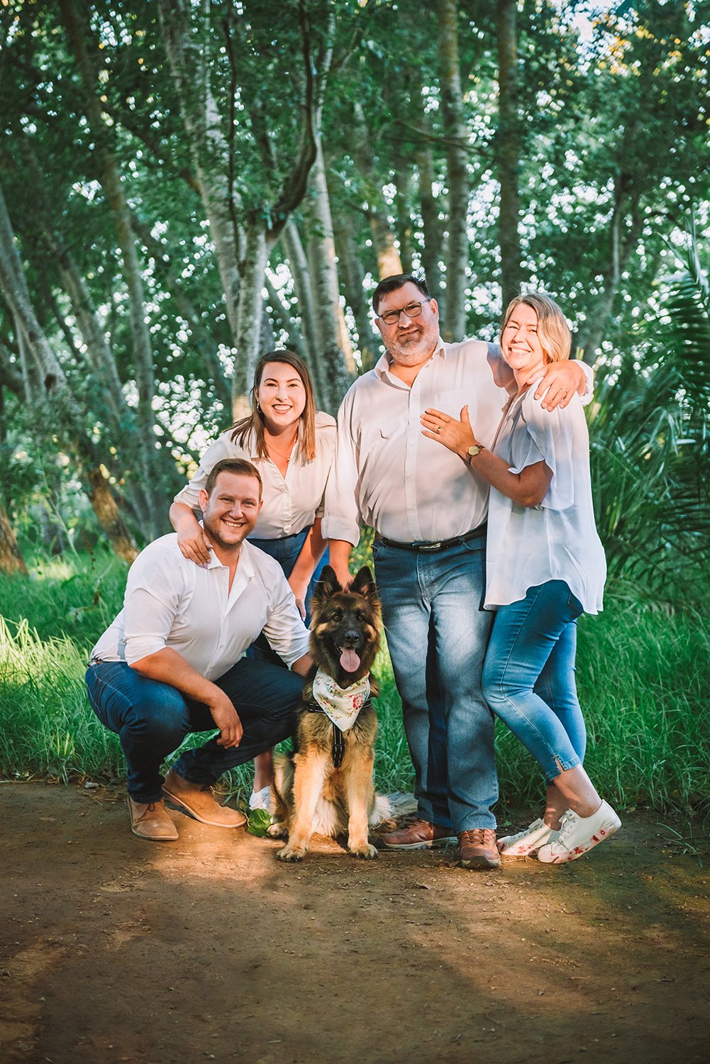 Family of husband, wife and wife's parents. All wearing white tops and blue jeans. Young German Shepherd Dog sits in the middle wearing a bandana.