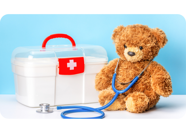Brown teddy bear with blue stethoscope sitting next to white box with a white cross on a red backgroud, indicating that it is a first aid box