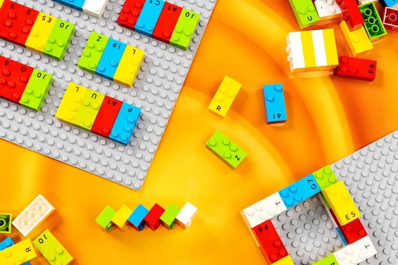 Peças de lego com as cores, vermelho, azul claro, verde claro, amarelo e cinza. Elas são adaptadas com braille