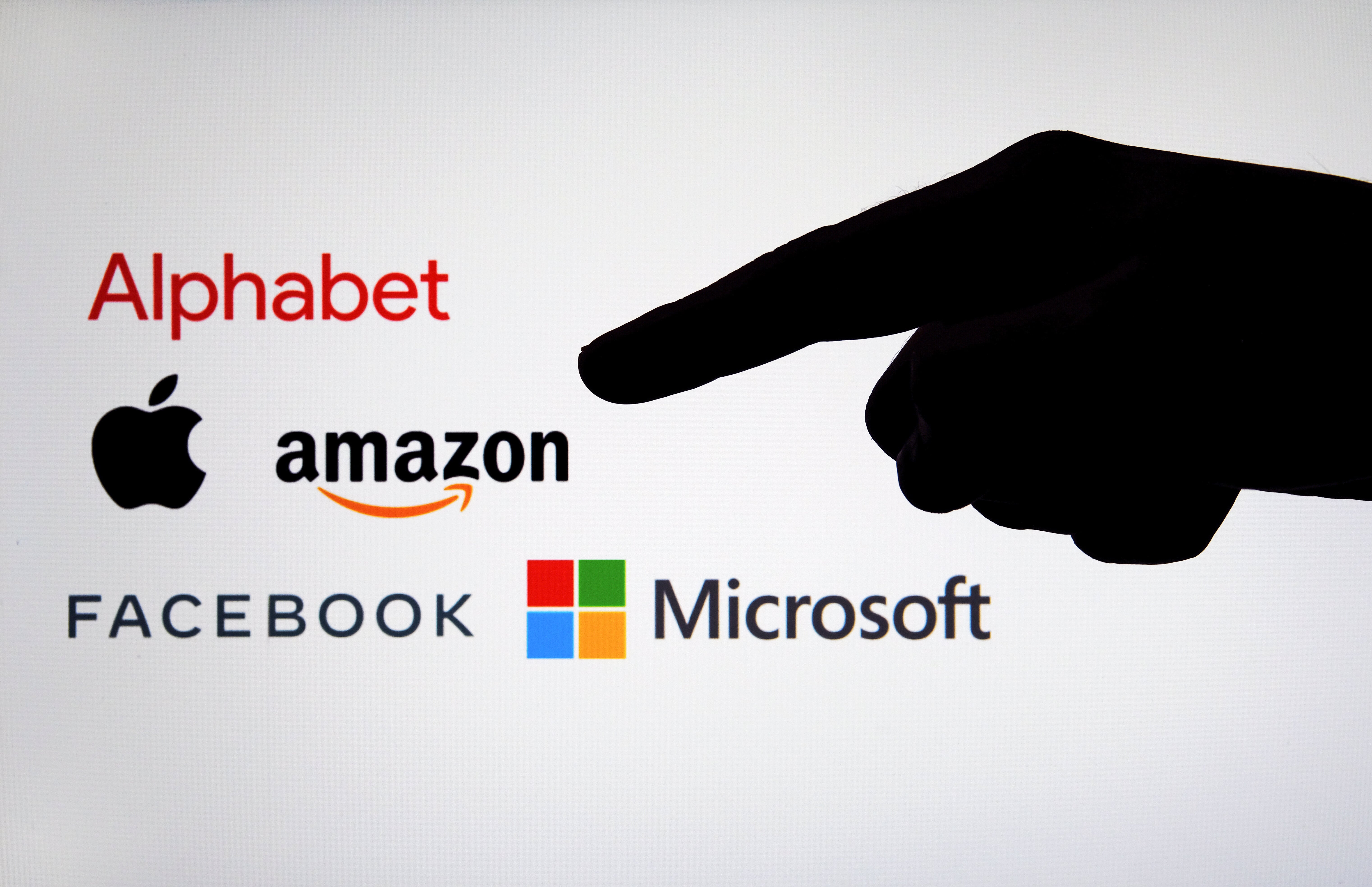 Big Tech company logos: Alphabet, Amazon, Apple, Facebook and Microsoft on a blurred screen and a silhouette of the hand. Concept for big tech regulation and antitrust.