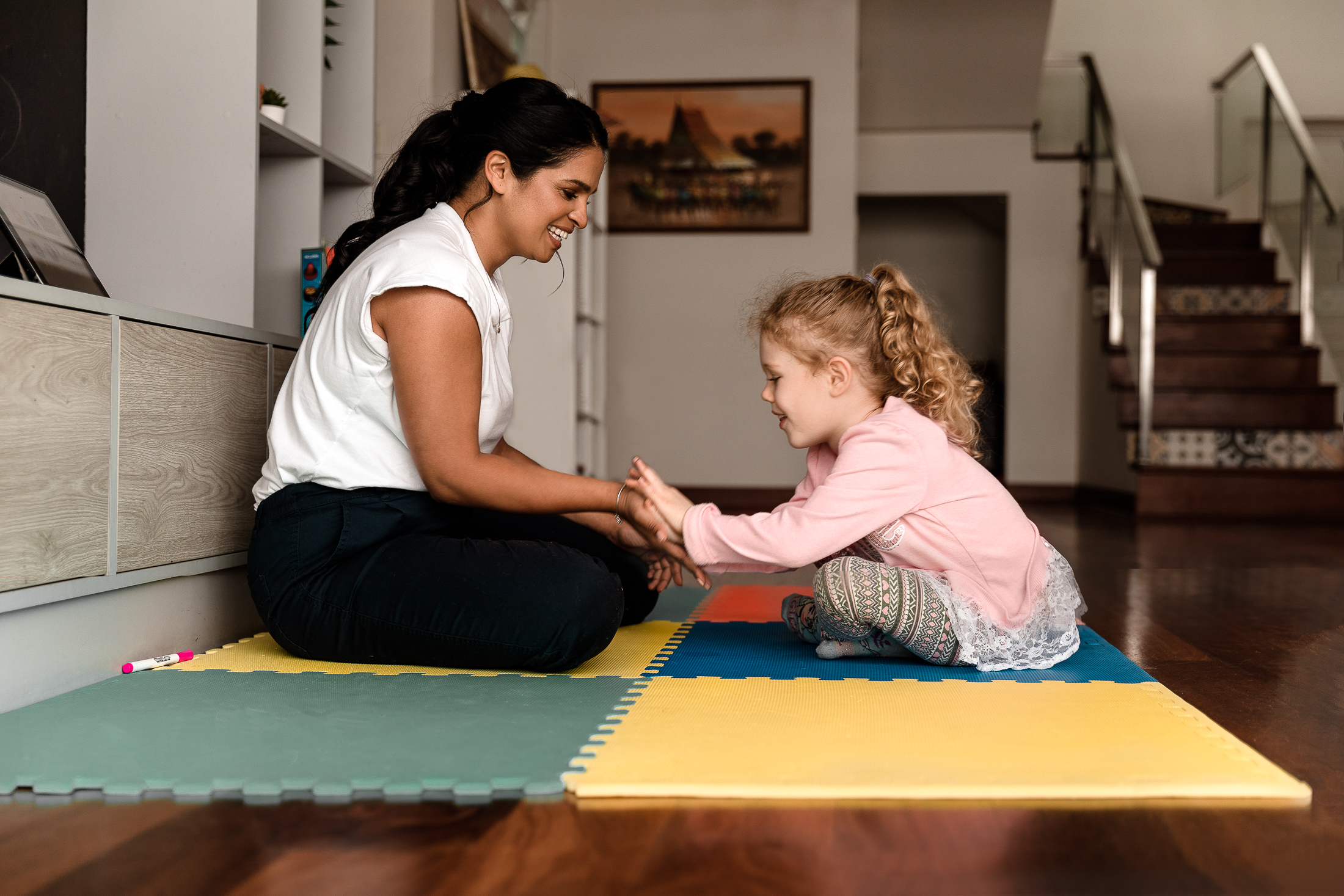 A speech and language therapist is sat face to face on a floor mat playing with a girl. They are smiling and doing a 'low ten'.