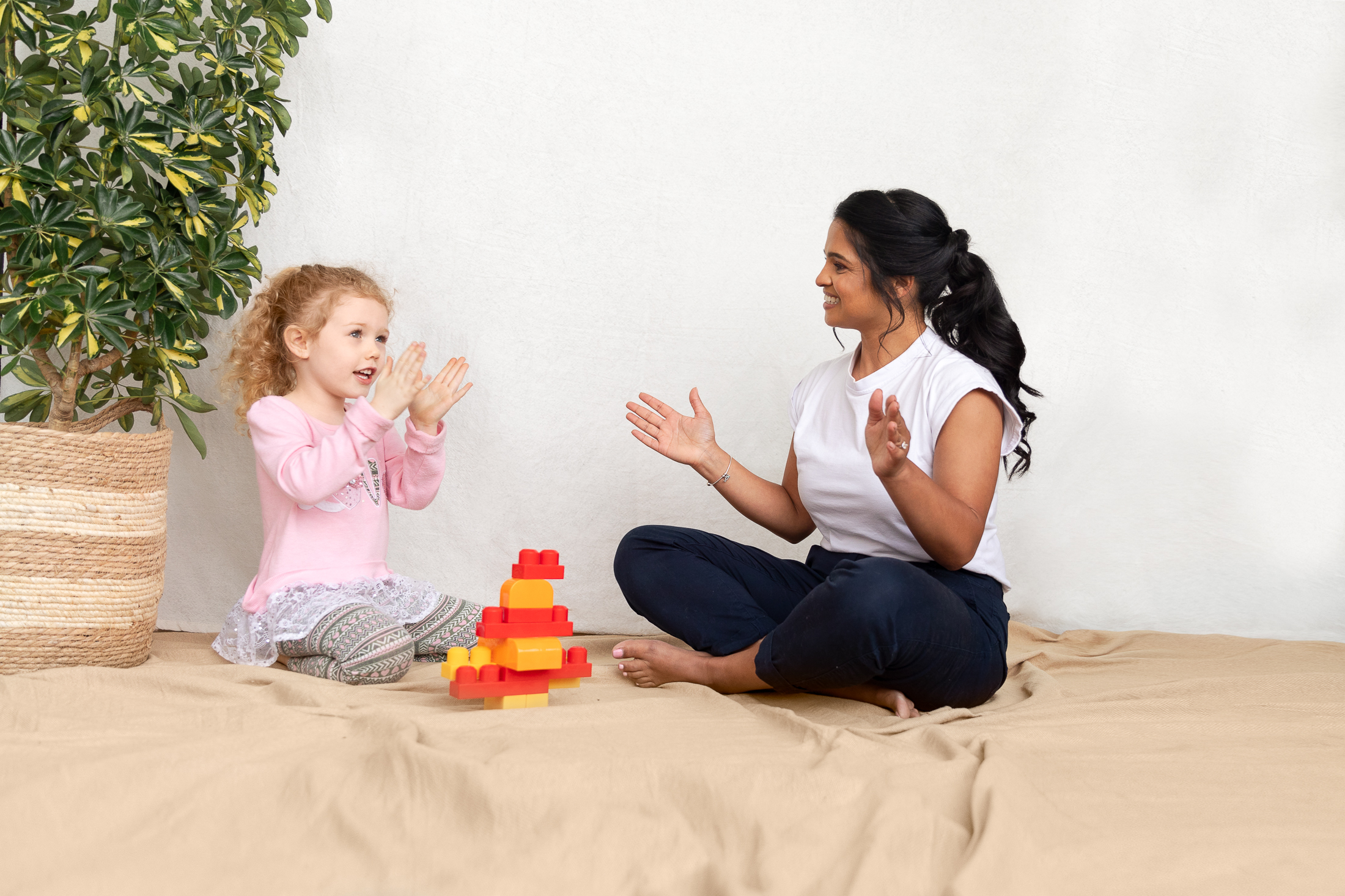 A speech and language therapist is sat face to face on a floor mat playing with a girl and clapping as they play with blocks..