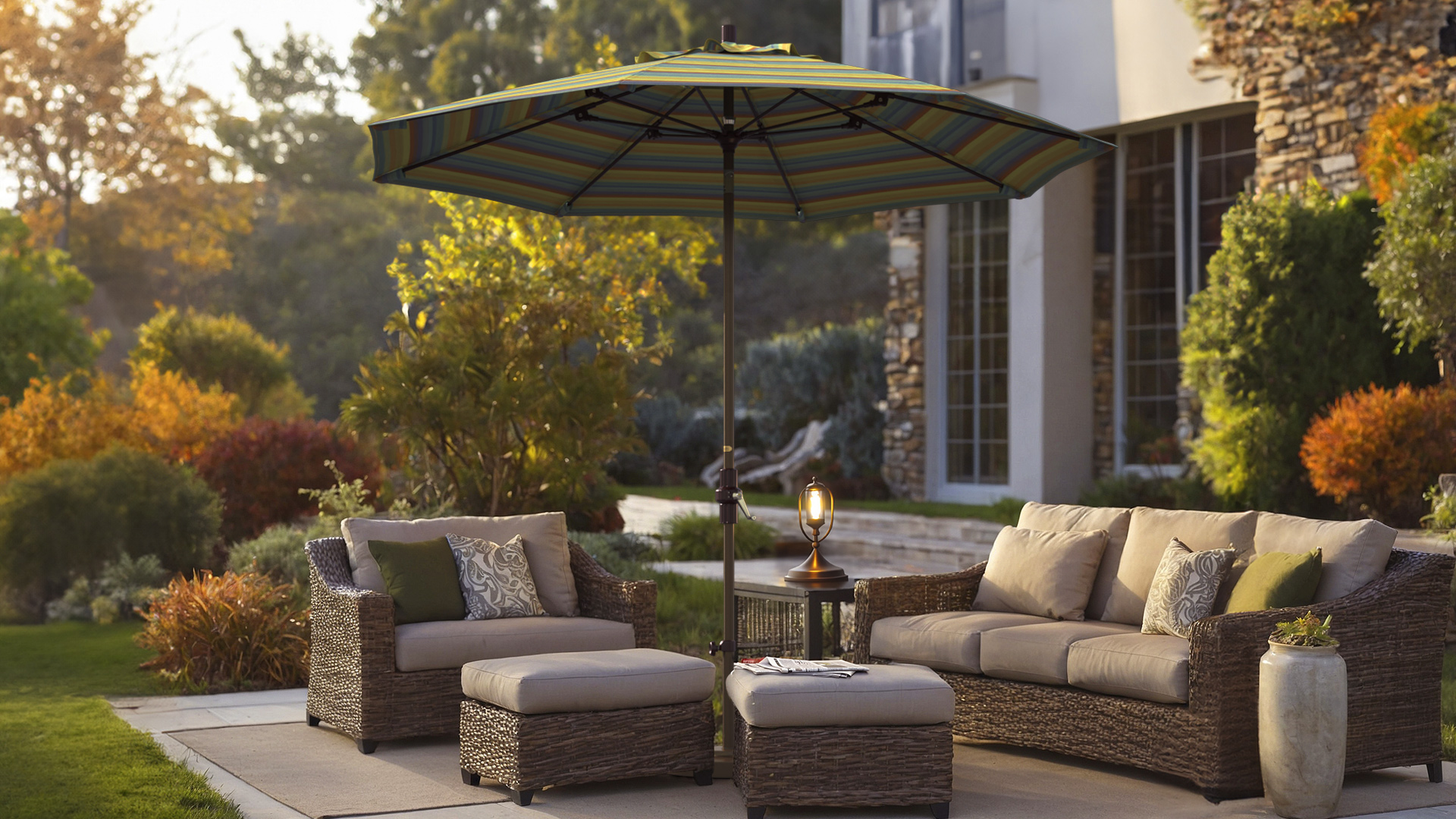 Cozy outdoor seating area with a California Umbrella Sun Master patio umbrella featuring a Sunbrella striped canopy, set against a lush autumn garden with warm, inviting colors. The wicker furniture provides a comfortable and stylish outdoor retreat.