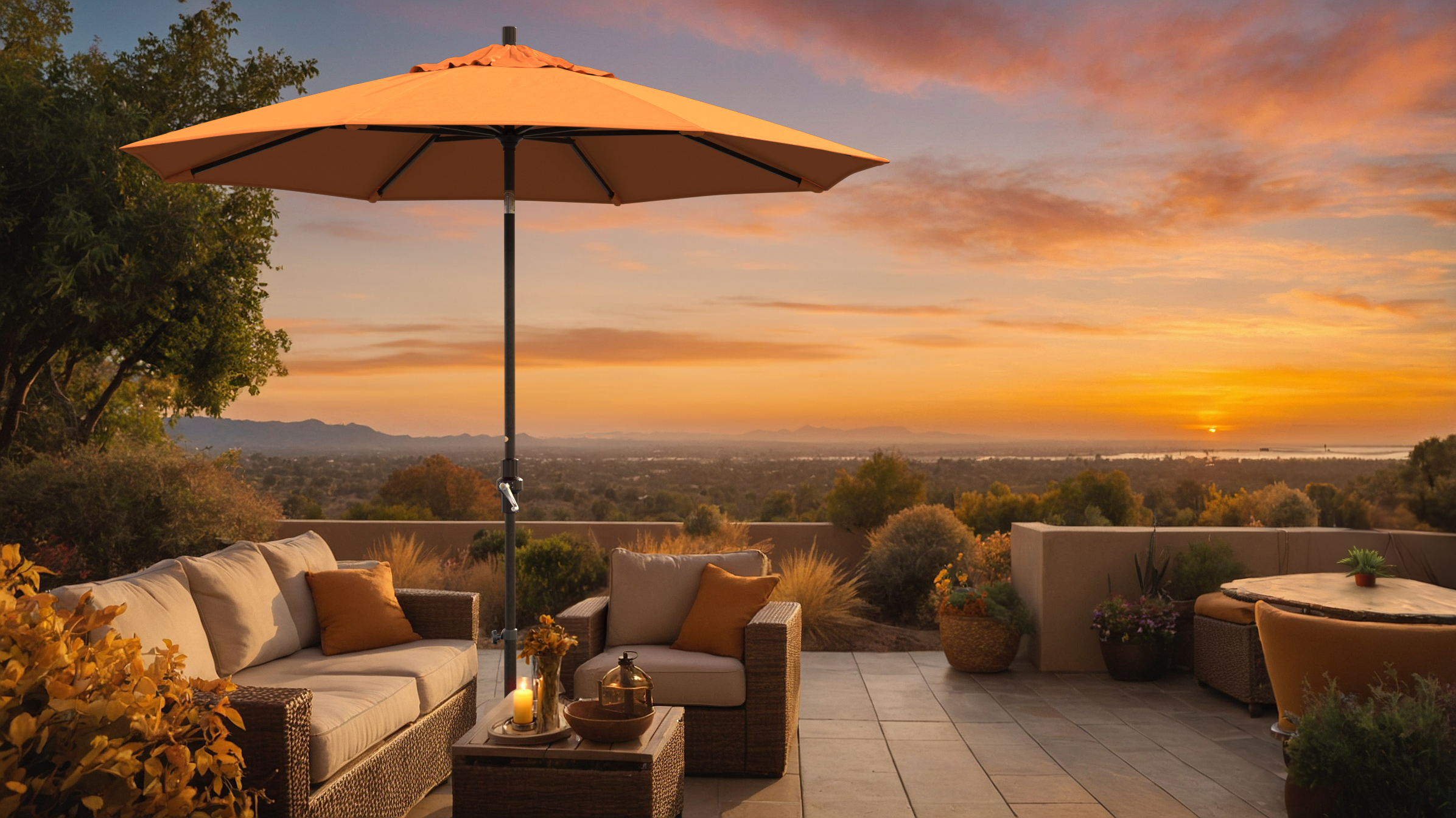 A peaceful patio scene at sunset featuring the Golden State Series California Umbrella with Sunbrella fabric in a warm tangerine color. The umbrella shades a cozy wicker seating area adorned with autumn-inspired cushions and candles, set against a stunning view of rolling hills and a glowing horizon.