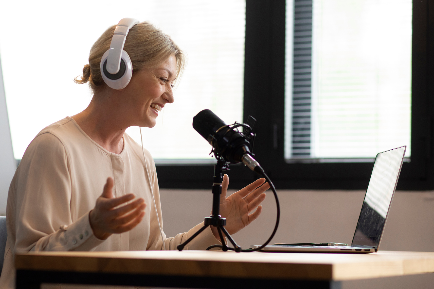 A woman wearing white headphones and a light blouse is speaking into a professional microphone while looking at her laptop. She is seated at a wooden desk in a modern, well-lit room with large windows in the background. She appears engaged and expressive, suggesting she is conducting or participating in an interview or podcast recording.
