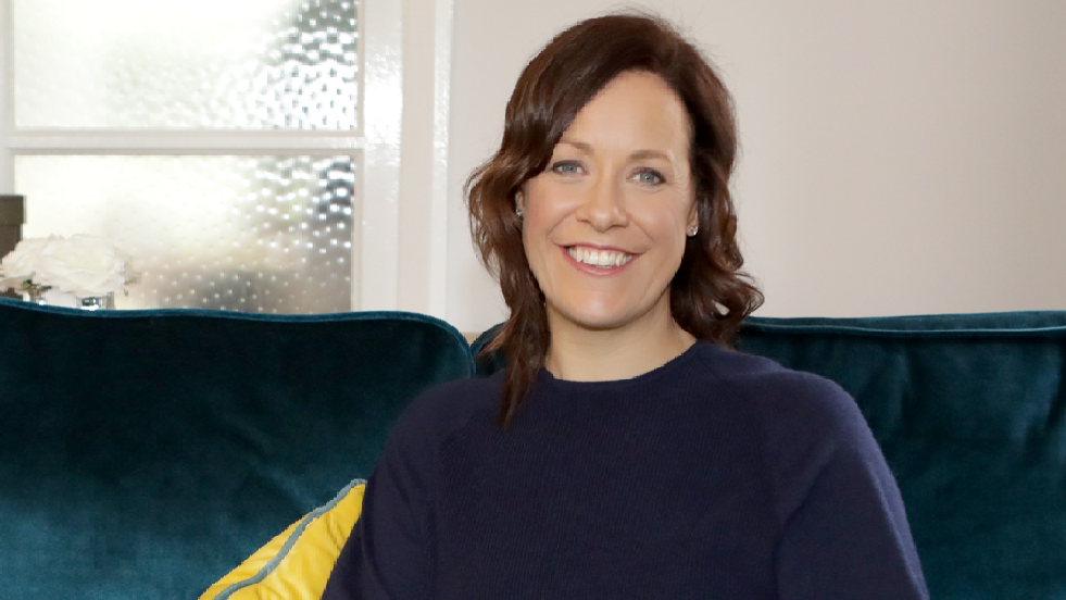 Woman smiling sat on a teal sofa wearing a navy blue jumper and jeans, holding a black notepad