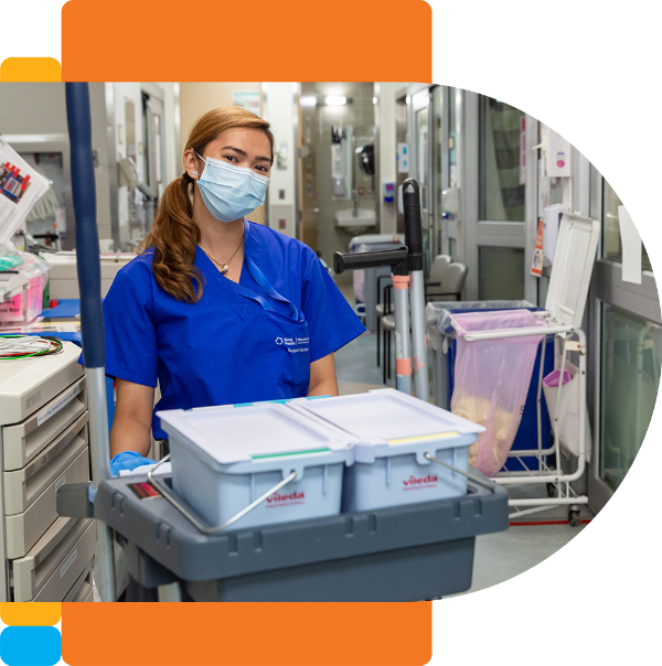 A nurse wearing a face mask and blue shirt stands behind a push cart holding containers. In the background there is a hospital hallway.