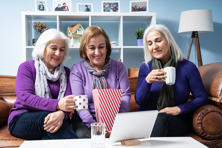three senior women sitting on a sofa and having a call on a tablet while sipping their coffee or tea