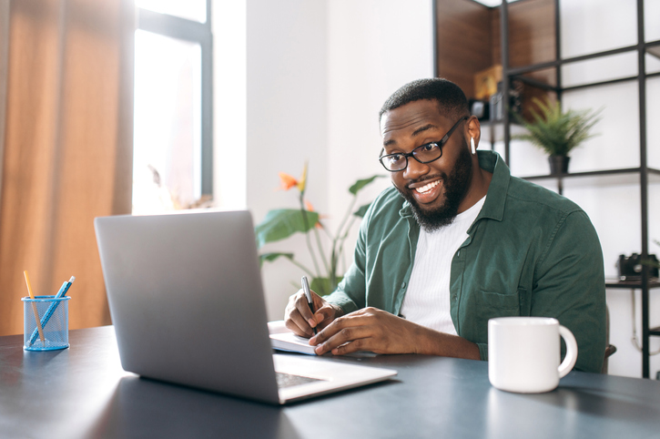African American smiling guy having fun during an online Italian session and taking notes on a notebook