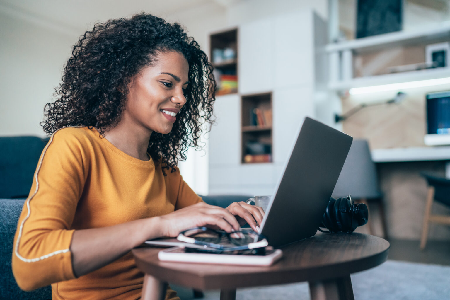 Woman smiling while using a laptop at home, with headphones on the table.