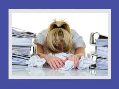 Female with blonde hair with head on desk holding scrumpled pieces of paper and ring binders either side of her head