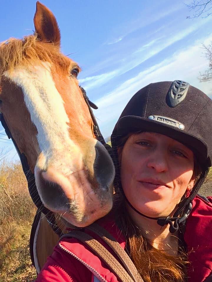 Image of a chestnut horse with a white blaze wearing a bridle without a noseband with its nose on Sophie's shoulder, a smilng female wearing a red jacket and black riding had.