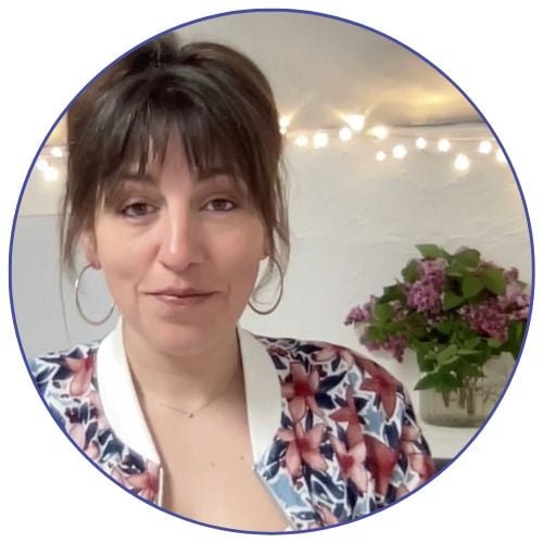 Image of Sophie within a circular frame. Female with dark hair tied up, large hoop earrings, a pink, blue and green floral jacket with a white collar and a fine necklace. Fairy lights and a vase of large flowers in the background.