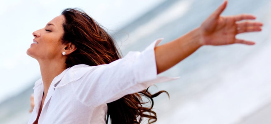 Side profile of tanned female with long dark hair and pearl earrings with her eyes closed arms raised upwards behing her looking care free, standing outside