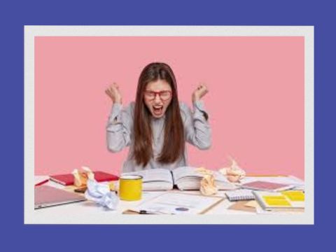 Female with long dark hair with a cluttered desk in front of her raising her fist, closing her eyes screaming, pink background