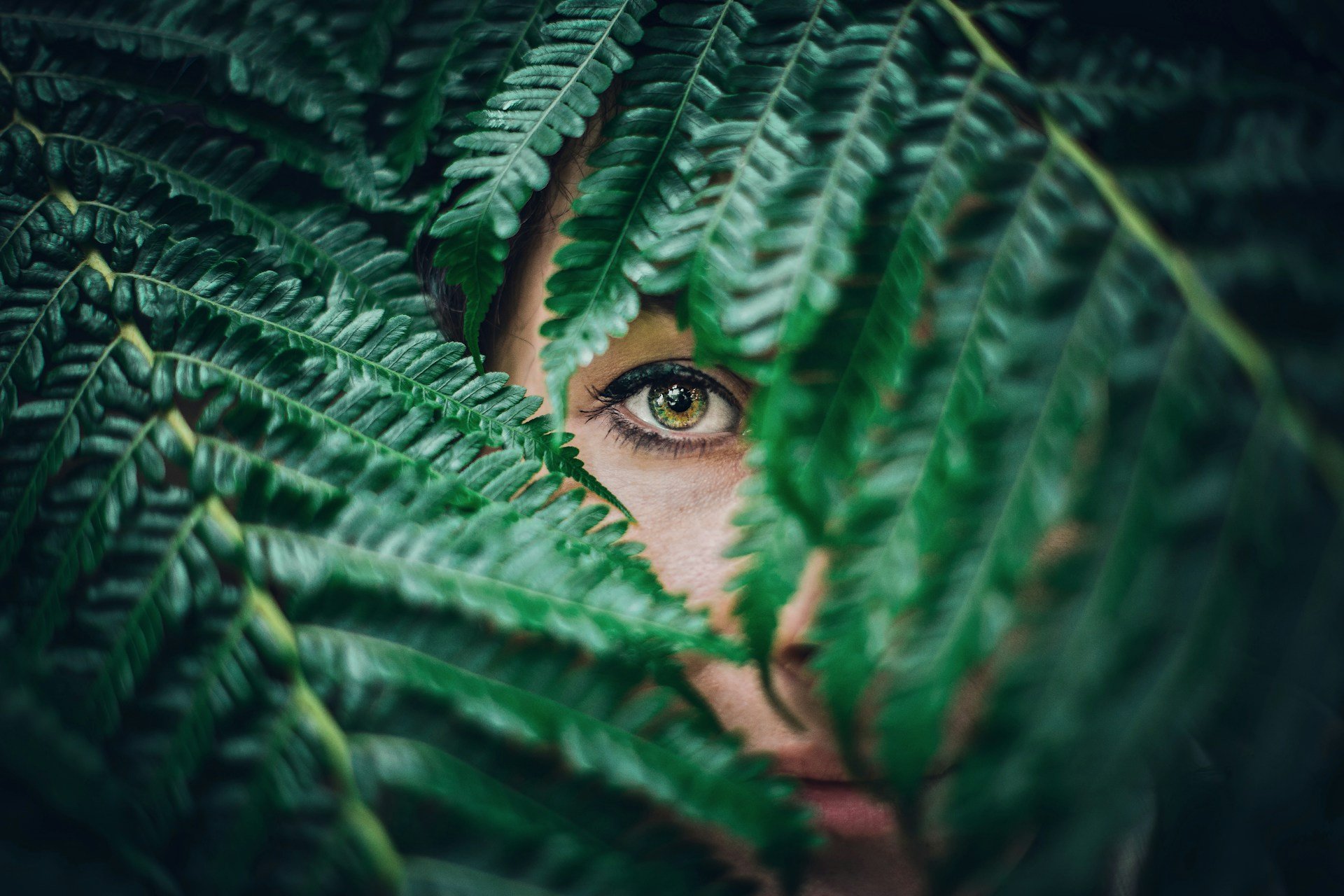 Woman's face partially hidden behind fern leaves, looking out with one eye