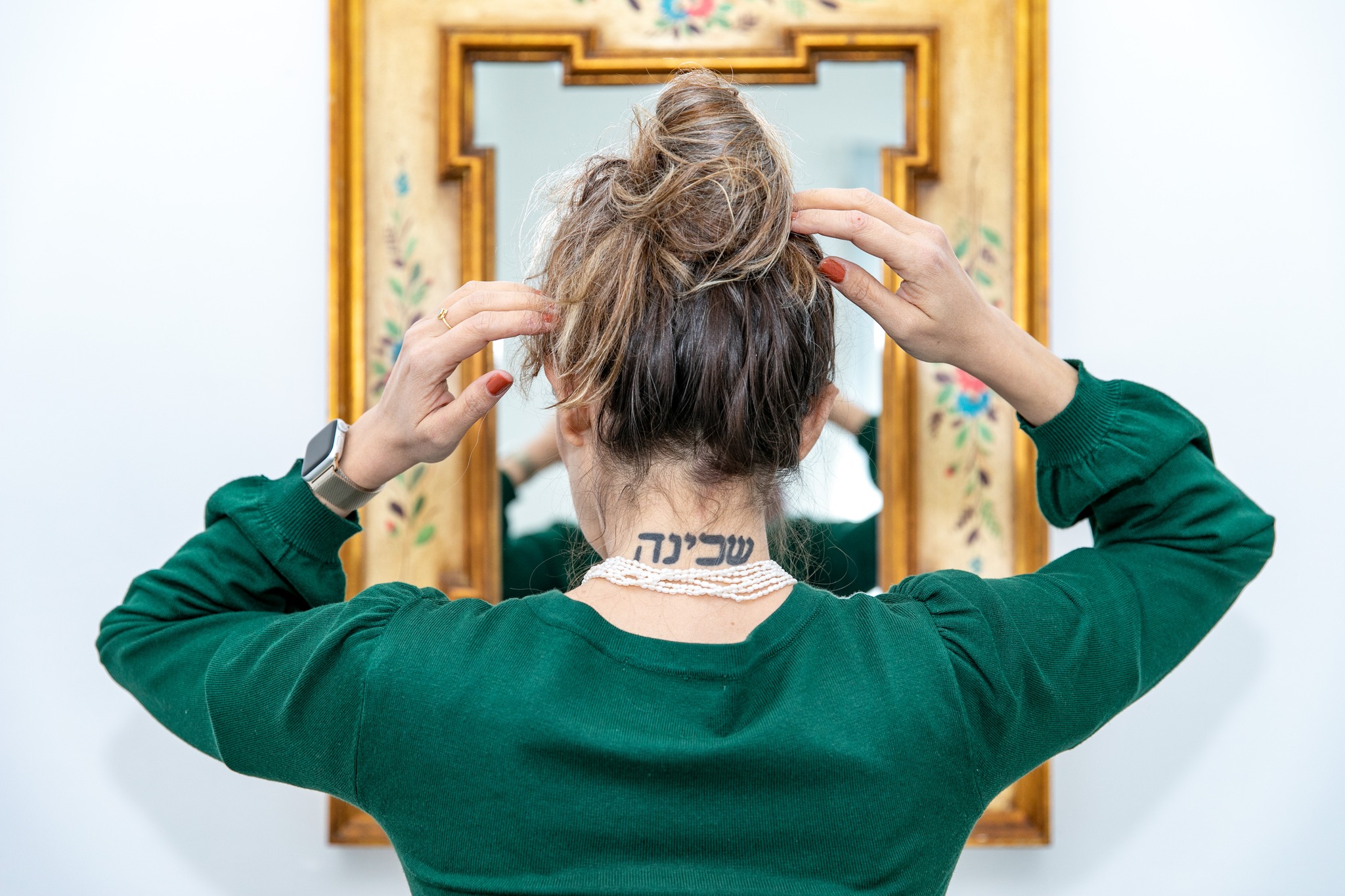 Foto di spalle di Emanuela Masia, si guarda allo specchio e si sistema i capelli, si vede un tatuaggio sul collo in aramaico o ebraico antico