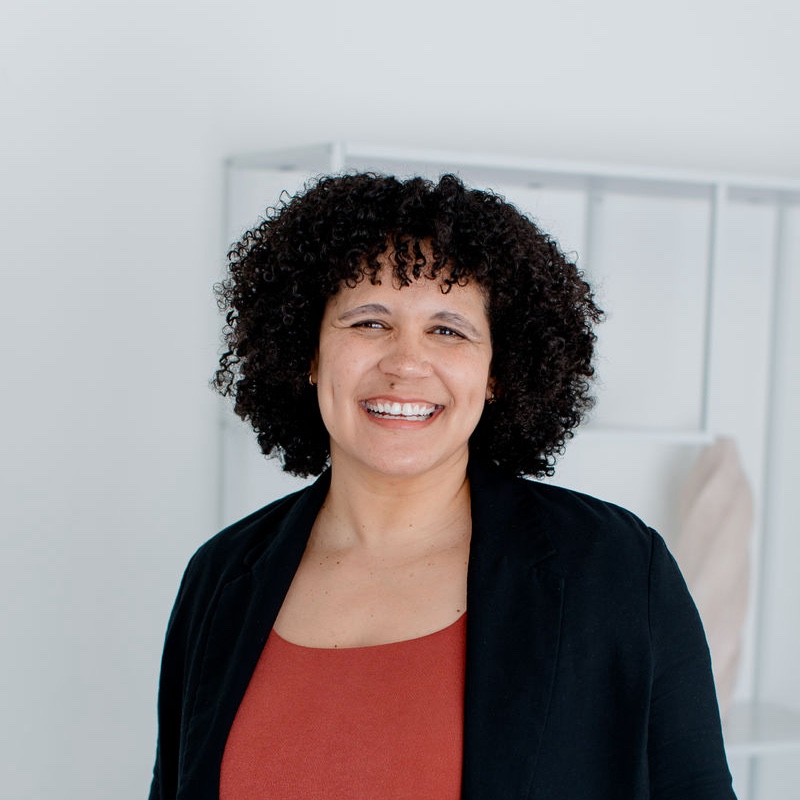 A woman wearing a red shirt and black blazer in front of a gray wall