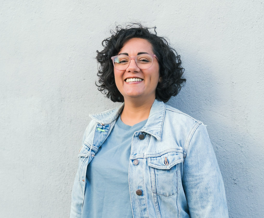 Sumi Fitzgerald, a brown person with a dark curly bob and pink glasses grinning