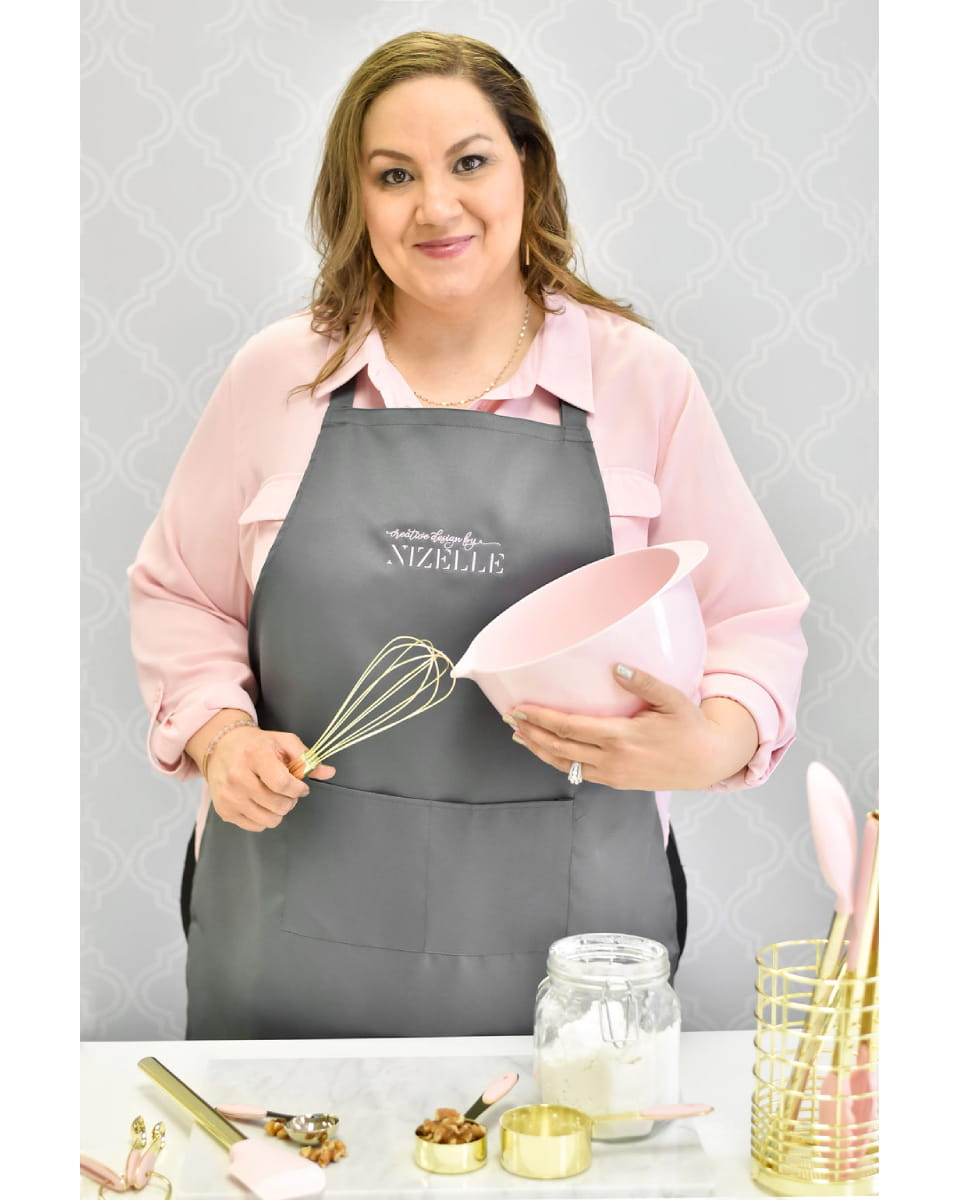 Nizelle, a professional cake designer in a grey apron, smiling while holding a pink mixing bowl and gold whisk. The scene is styled with elegant gold and pink baking tools on a marble counter.