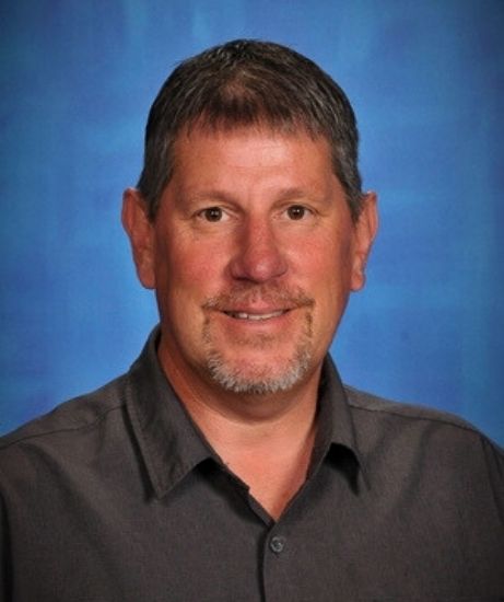 Portrait of Don Baribault against a blue studio backdrop. He is wearing a dark gray collared shirt and smiling gently.
