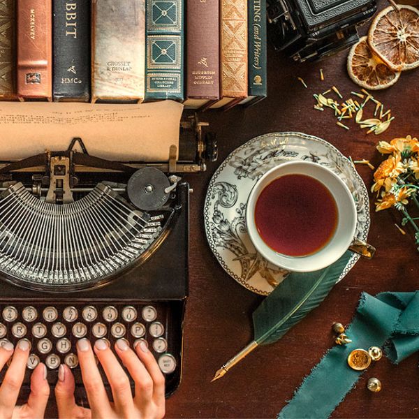 Author K.T. Jay typing on her typewriter with books, a cup of tea, a quill, and flowers on her desk.