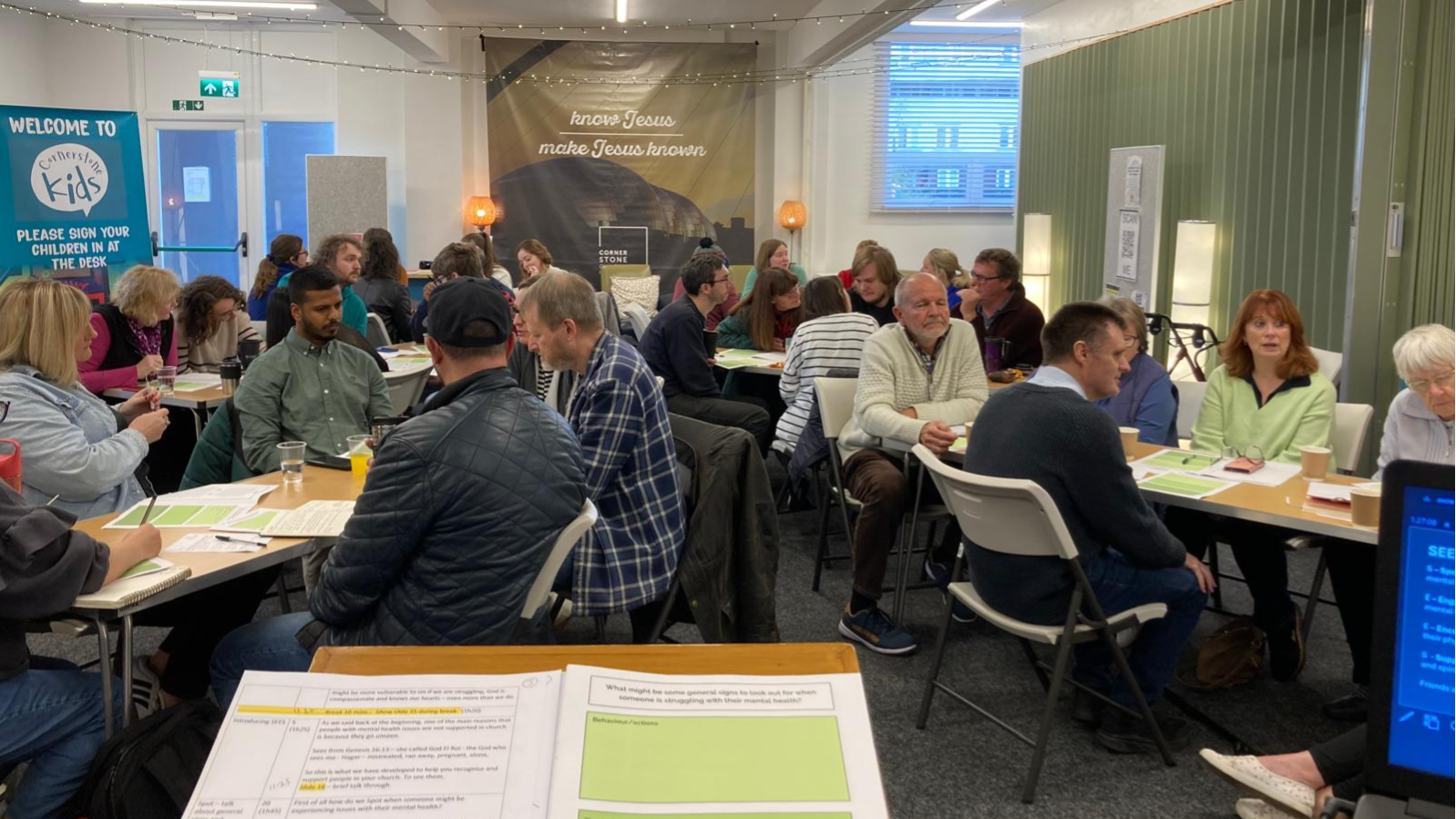 photo from one of our church mental health days showing about 20 people sitting around tables in groups in a church building. They have pens and paper in front of them and they are in discussion with each other. in the foreground of the picture is a lectern with notes on it.