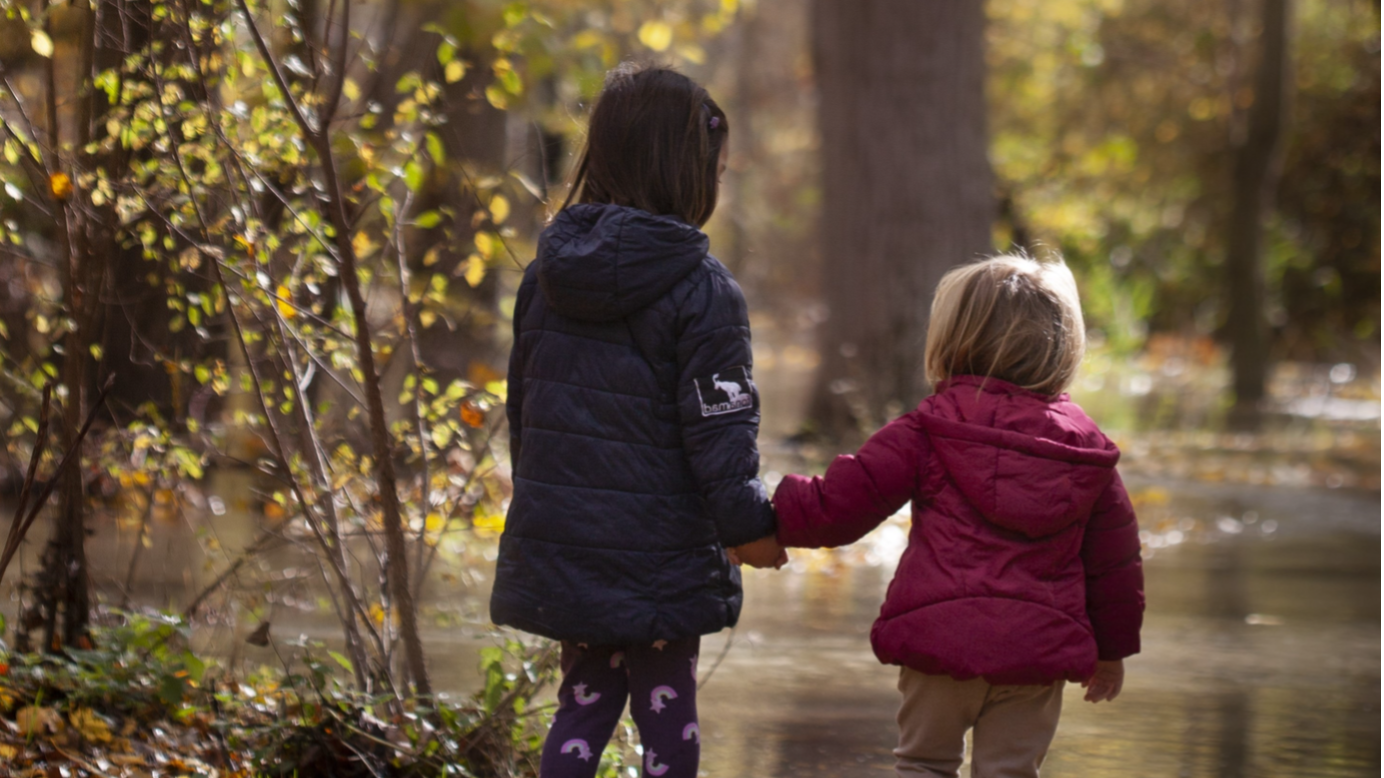 Two children in coats holding hands as they stand by a lake surrounded by trees