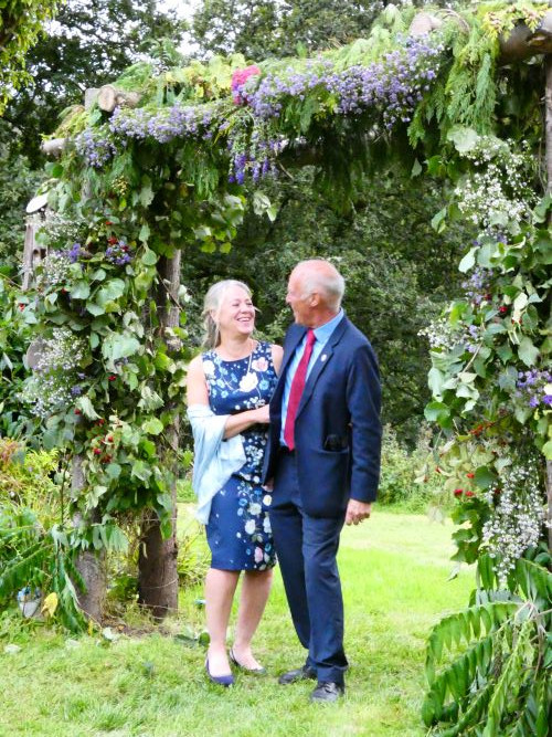 Kevin and Michelle in matching blue suit and blue shift dress standing beneath a floral pergola