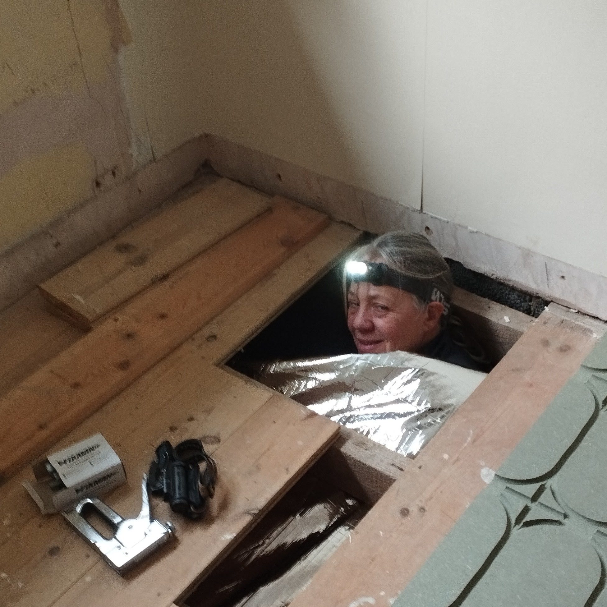 Michelle's head wearing a head torch is popping up from under the floorboads as she installs the underfloor insulation