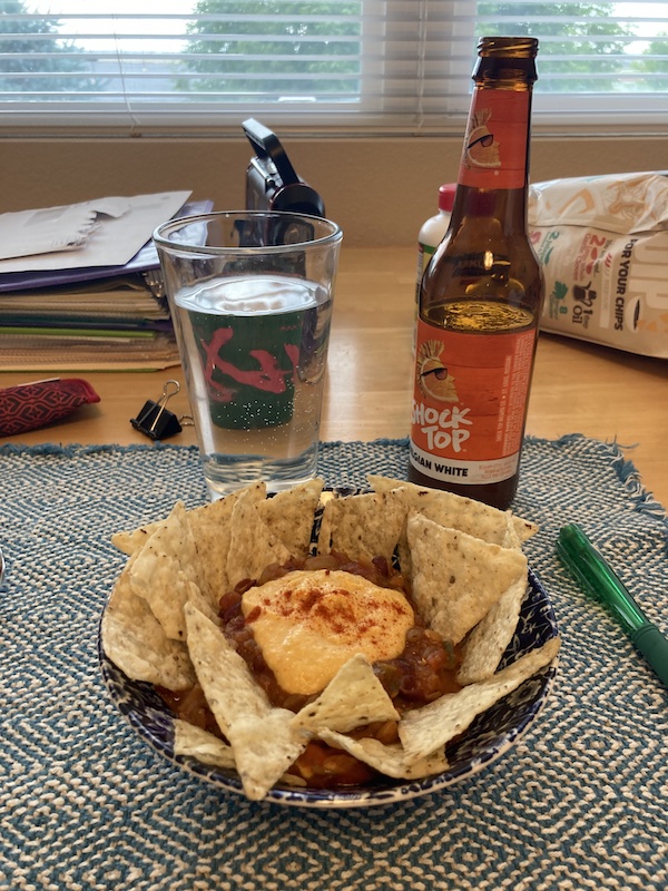 A plate of chili cheese nachos is on a plate in the forground. The golden-orange cheese is sprinkled with smoked paprika. Behind the plate is a bottle of Shock Top beer and a pint glass of the same. Behind the beer is a radio, reading glasses in a case, a binder clip and a binder with papers inside. Next to that is a fast food bag. Beyond this arrangement of nutritional bliss is a window looking out onto some trees.