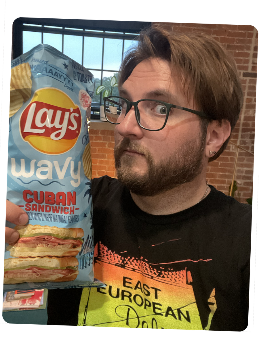 A late-30's white man with a beard and eyeglasses holding a bag of Lay's Wavy Cuban Sandwich flavor potato chips.