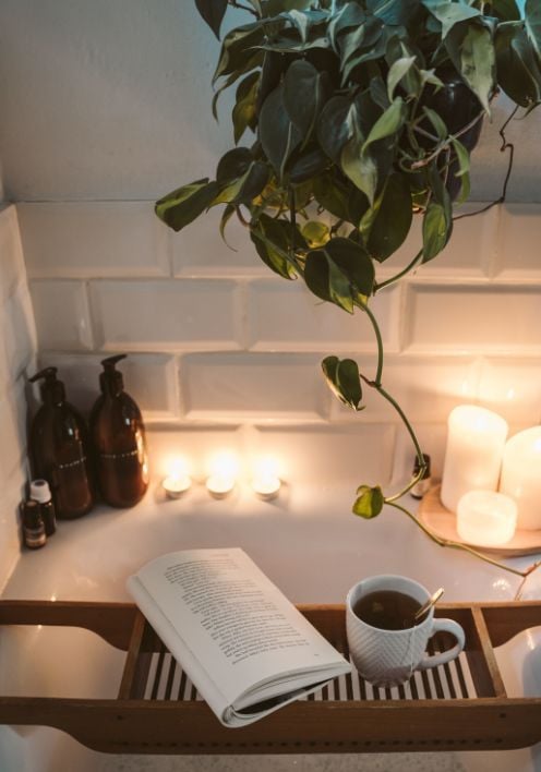 an image of an open romantic comedy book and a coffee on a tray in a relaxing bathtub with candles and a green plant 