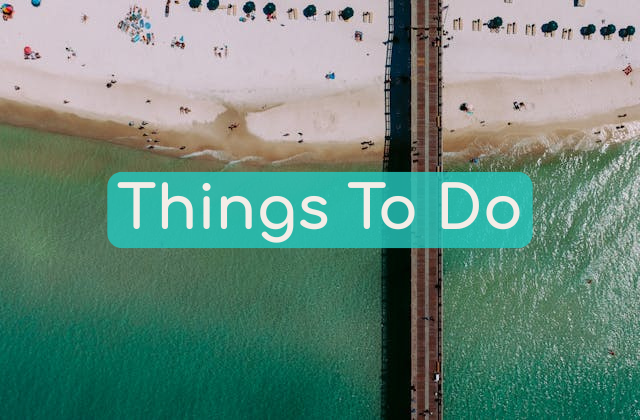 Beautiful aerial picture of the beach in Panama City Beach, FL on the Florida Emerald Coast which includes the soft white sand, calming turquoise waters, and a few beach chairs and people scattered along the sandy beachside in view as well as the pier jutting out into the water, presented as the image for the Near Me Discovery blog content hub image for Things To Do near me and near you, including local businesses and small businesses such as local products and local services, bars, restaurants, shops, entertainment, shows, and other local attractions